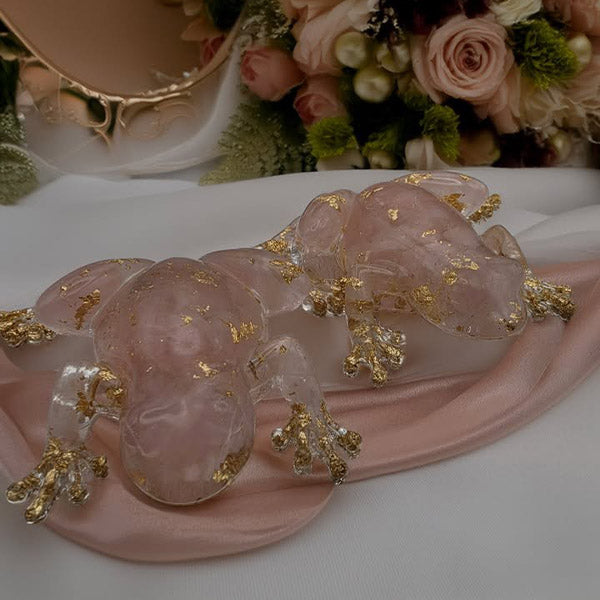 A pair of rose quartz crystal frog-decorated with gold Leaf and gold branch details, resting on a pink satin ribbon.