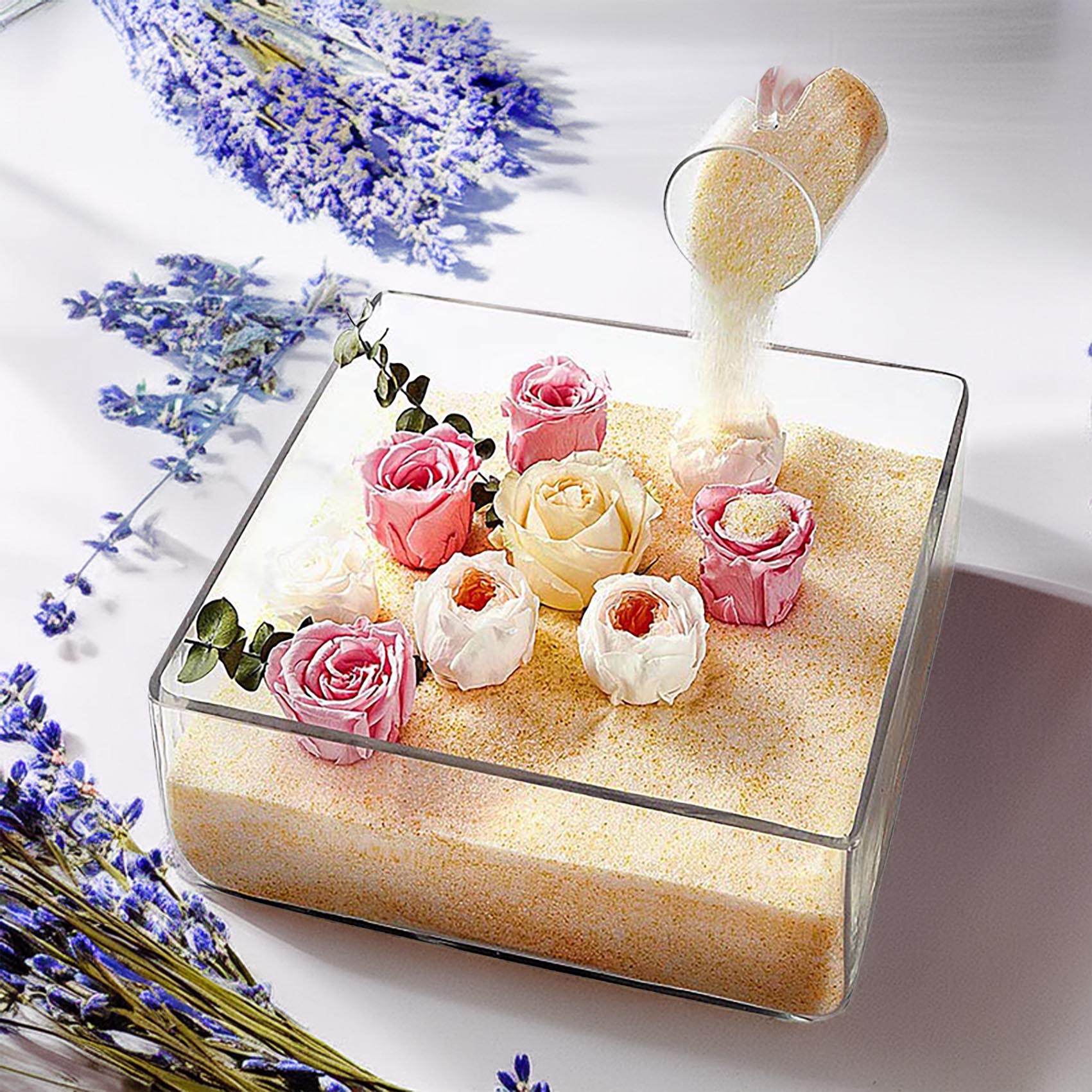 A square clear glass container filled with pale golden sand or sugar, decorated with several small artificial roses in pink, cream, and white, with more flowers scattered around it and a small glass pouring sand into the container.